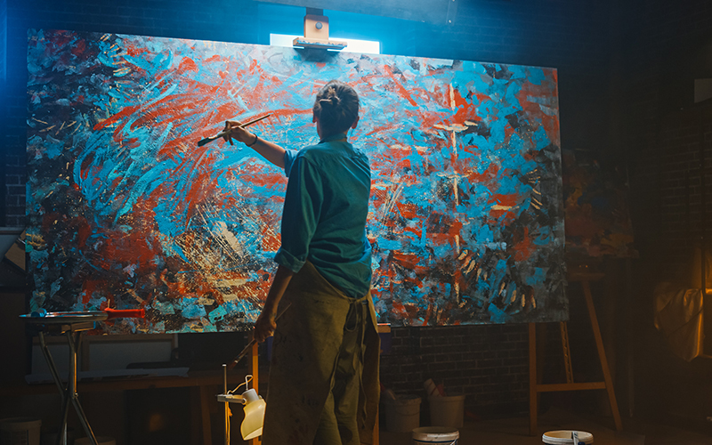 Talented Female Artist Works on Abstract Oil Painting, Using Paint Brush She Creates Modern Masterpiece. Dark and Messy Creative Studio where Large Canvas Stands on Easel Illuminated.